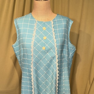 May include: A sleeveless, vintage-style dress in a light blue hue with a white grid pattern. The dress features a white lace trim down the center and three round, yellow buttons. The dress is displayed on a mannequin.