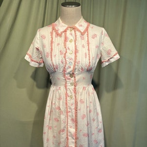 May include: A white cotton dress with a pink floral print and pink lace trim. The dress has a button-up front and short sleeves.