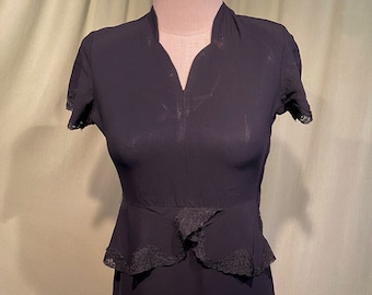 May include: A vintage black dress with short sleeves and a V-neckline. The dress features a peplum waist with lace detailing and a knee-length skirt. The dress is displayed on a mannequin.