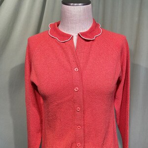 May include: A coral pink cardigan with a white scalloped collar and button closure. The cardigan is made of a textured fabric.
