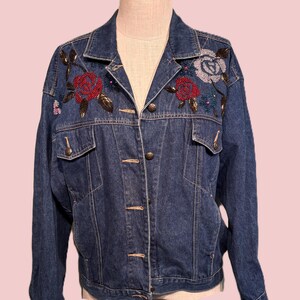 May include: A dark blue denim jacket with floral embroidery. The jacket features a classic collar, button-down closure, and two chest pockets. The floral embroidery includes red, blue, and white roses with gold-colored leaves.