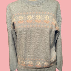 May include: A light blue sweater with a crew neck and long sleeves. The sweater features a floral pattern in white and peach across the chest and near the hem. The knitwear is displayed on a mannequin.