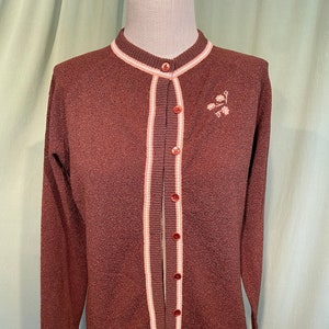May include: A brown cardigan sweater with a round neckline and long sleeves. The sweater has a white and peach stripe down the front and around the neckline. It features a small floral embroidery on the upper left chest and is buttoned up.