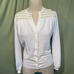 May include: White cardigan sweater with a V-neck and button closure. The sweater has a textured knit and a delicate yellow and white trim around the neckline.