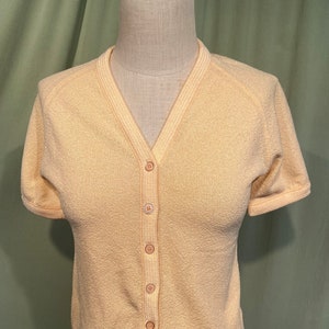 May include: A pale yellow short-sleeved cardigan with a V-neck and button closure. The cardigan has a ribbed texture and a slightly cropped length.