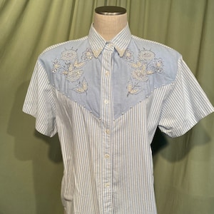 Puede incluir: Una camisa de manga corta de botones azul claro con rayas blancas y bordado floral de perlas en los hombros.