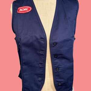 May include: A navy blue vest with a V-neck and button closure. The vest features a red and white "ACME" patch on the chest. The vest is displayed on a mannequin against a pink background.
