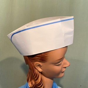 以下が含まれることがあります： 青い縁取りのある白い紙製のシェフハット。茶色の髪のマネキンヘッドに被せられています。
