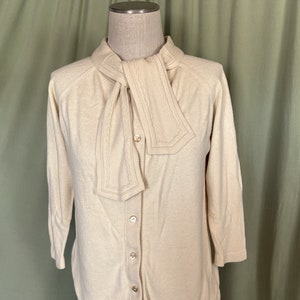 May include: A cream-colored cardigan with a long, tied collar and button closure. The collar has a decorative embroidered trim.