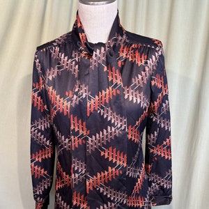 Op de afbeelding: Een zwarte blouse met lange mouwen met een geometrisch patroon in tinten bruin, oranje en wit. De blouse heeft een hoge kraag en een strik om de hals.