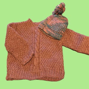 May include: A knitted sweater and matching beanie set. The sweater is a textured, reddish-brown color with a cable knit detail on the front. The beanie is striped with shades of teal, orange, and brown, and has a pom-pom on top.