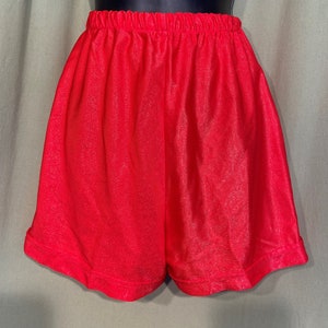 May include: Red athletic shorts with a shiny, textured fabric. The shorts have a wide elastic waistband and a relaxed fit.