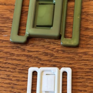 May include: Two plastic belt buckles. One is olive green, and the other is white. Each buckle has a rectangular center piece with two rectangular loops on either side.
