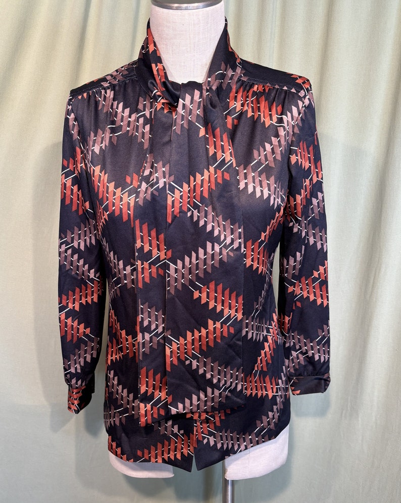Op de afbeelding: Een zwarte blouse met lange mouwen met een geometrisch patroon in tinten bruin, oranje en wit. De blouse heeft een hoge kraag en een strik om de hals.