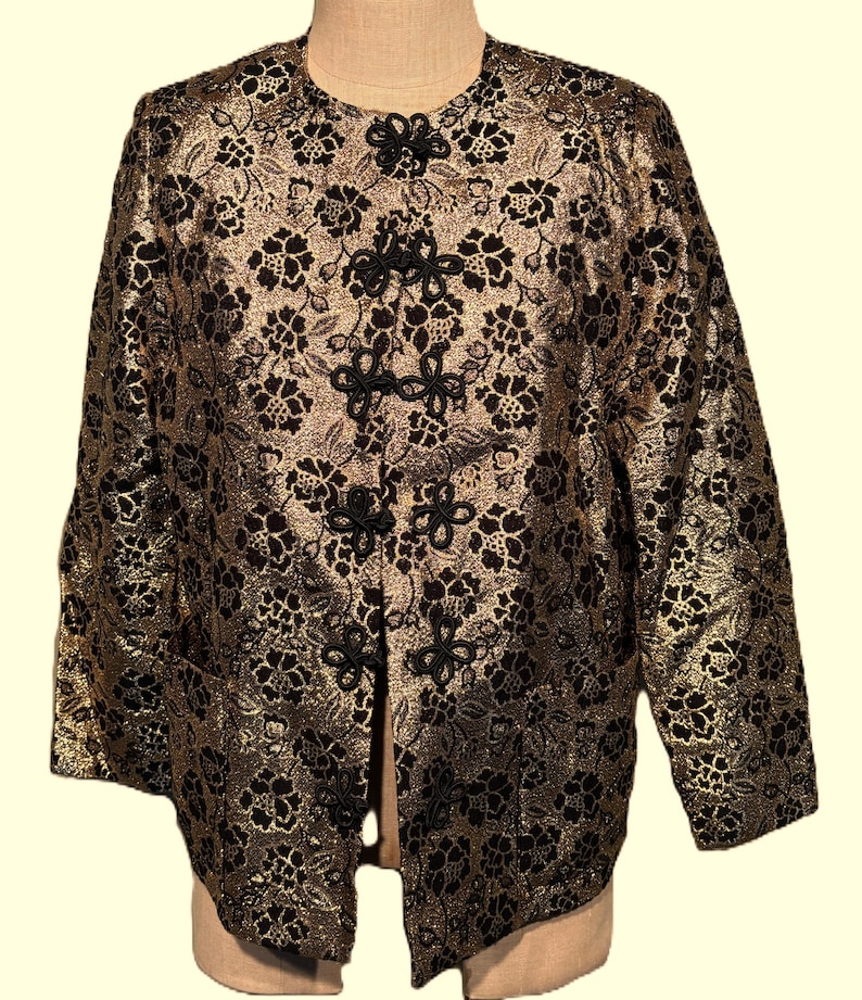 May include: A gold and black brocade jacket with a mandarin collar and decorative black buttons. The jacket has a floral pattern and a long sleeve.