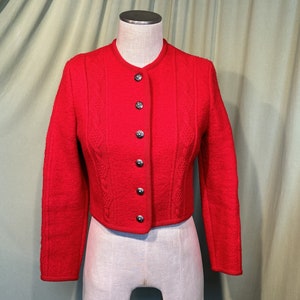 Op de afbeelding: Een rood gebreid jasje met een hoge hals en een rij zilveren knopen aan de voorkant. Het jasje heeft een getailleerde lengte en een kabelbreisel.