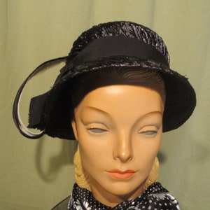 Może przedstawiać: Czarny aksamitny kapelusz z szerokim rondem i dużą kokardą z boku. Kapelusz znajduje się na głowie manekina.