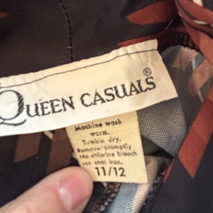 Op de afbeelding: Een wit label met de tekst "QUEEN CASUALS" in zwarte letters en een kroonsymbool. Het label bevat ook wasinstructies voor machinewas, drogen in de droger, snel verwijderen, geen chloorbleekmiddel en strijken op lage temperatuur. Het label heeft een maat van 11/12.