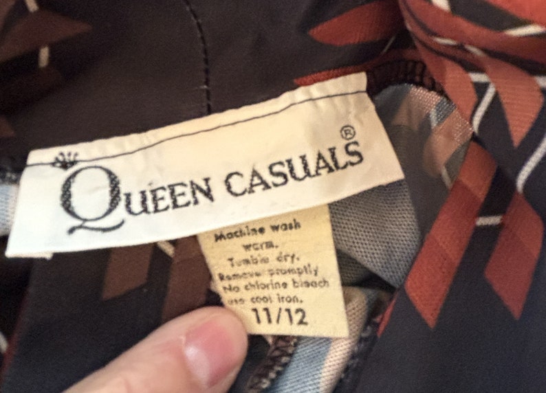 Op de afbeelding: Een wit label met de tekst "QUEEN CASUALS" in zwarte letters en een kroonsymbool. Het label bevat ook wasinstructies voor machinewas, drogen in de droger, snel verwijderen, geen chloorbleekmiddel en strijken op lage temperatuur. Het label heeft een maat van 11/12.
