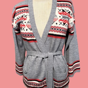 May include: Gray cardigan sweater with a wrap-around belt. The sweater features a V-neck and pockets. The cuffs and upper chest have a patterned design in red, black, and white. A vintage style garment.