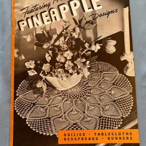 Op de afbeelding: Een vintage haakpatroonboek met 14 nieuwe ananasontwerpen. De omslag toont een close-up van een gehaakt doily met een boeket bloemen in een witte vaas. De titel van het boek is "Featuring 14 New Pineapple Designs". Het boek is getiteld "Clark's O.N.T. J&P Coats Book No. 230 Price 10 Cents".