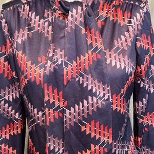Op de afbeelding: Een blouse met lange mouwen met een geometrisch patroon in tinten rood, roze en wit op een zwarte achtergrond. De blouse heeft een strik aan de hals.