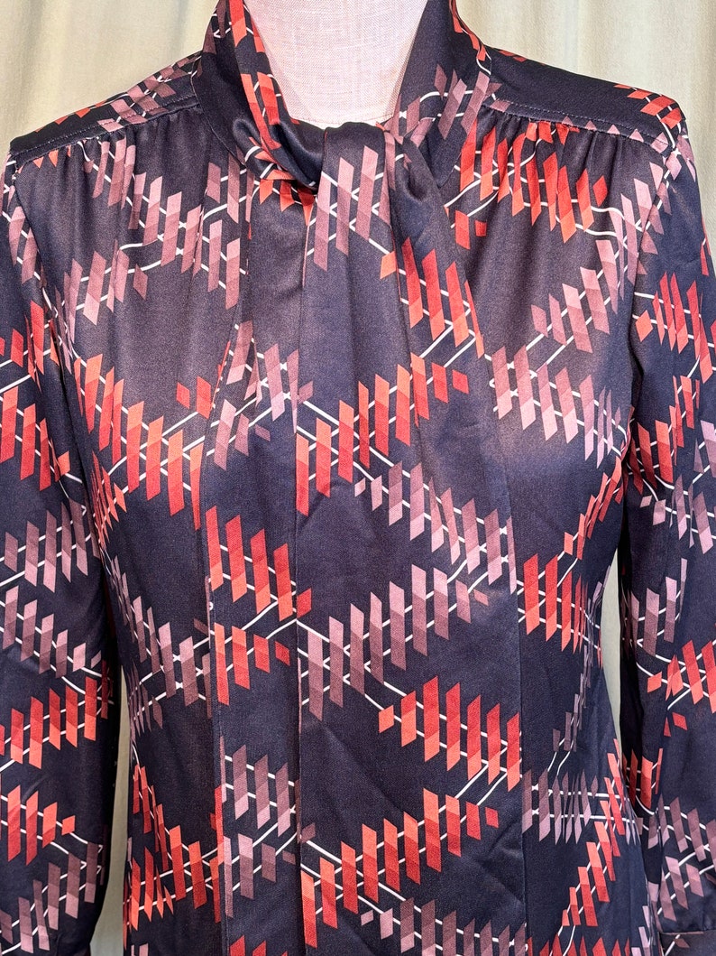 Op de afbeelding: Een blouse met lange mouwen met een geometrisch patroon in tinten rood, roze en wit op een zwarte achtergrond. De blouse heeft een strik aan de hals.