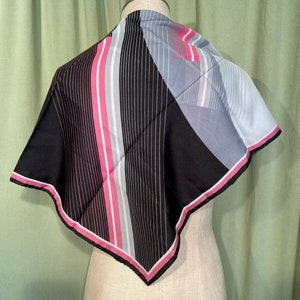 May include: A black, pink and grey patterned silk scarf with a geometric design. The scarf is folded into a triangle and displayed on a mannequin.
