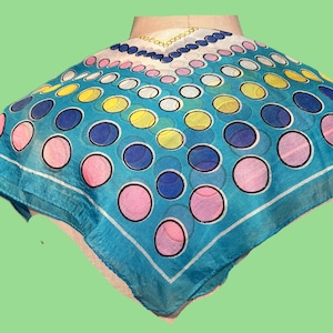 May include: A square scarf featuring a vibrant turquoise background with a repeating pattern of polka dots in shades of pink, yellow, and dark blue. The scarf has a white border and a decorative chain-link graphic along the top edge.