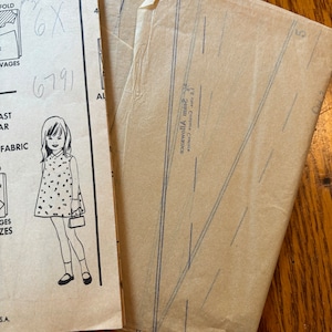 May include: Vintage sewing pattern with a drawing of a girl in a dress. The pattern is size 6X, with the number 6791 written on it. The pattern is folded and has instructions and measurements.