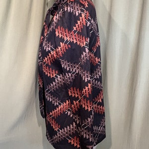 Op de afbeelding: Een zwart, lang mouwig blouse met een geometrisch patroon in tinten van bruin, oranje en wit. De blouse heeft een strik aan de hals.