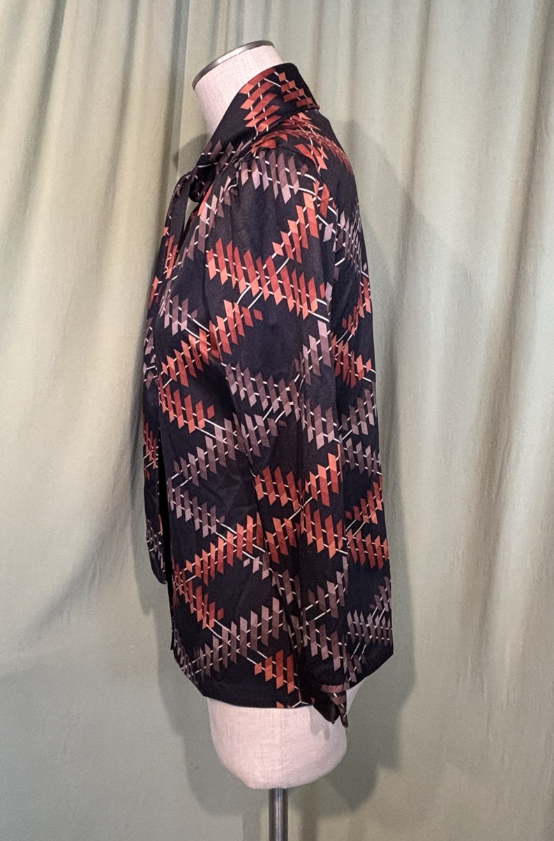 Op de afbeelding: Een zwart, lang mouwig blouse met een geometrisch patroon in tinten van bruin, oranje en wit. De blouse heeft een strik aan de hals.