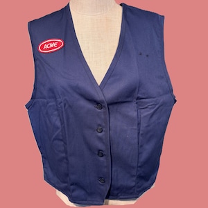 May include: A navy blue vest with a V-neck and button-up front. The vest features a red and white "ACME" patch on the chest. The vest is displayed on a mannequin against a pink background.