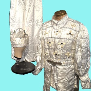 May include: A vintage cream-colored costume set, including a jacket, pants, and hat. The jacket features decorative shell accents and silver trim. The pants and hat are made of a matching satin-like fabric. The costume is displayed on a mannequin.