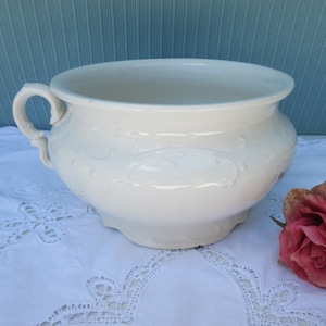 May include: White ceramic chamber pot with an ornate design. The pot has a single handle and a wide, round opening.