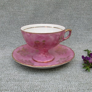 May include: A pink teacup and saucer set with gold trim and floral designs. The cup has a delicate handle and a rounded shape. The saucer has a matching floral design.