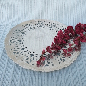 May include: A silver, decorative, round serving tray with a delicate, lacy design. A sprig of dried, burgundy-colored flowers rests on the tray.