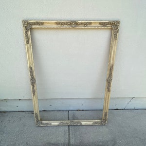 May include: A large, ornate, gold-colored picture frame with a decorative design. The frame is sitting on a concrete surface.