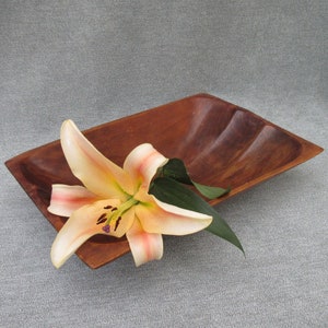 May include: A wooden serving bowl with a wavy edge, featuring a single peach-colored lily with green leaves.