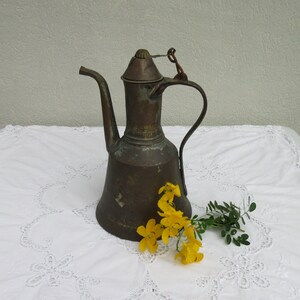 May include: A vintage copper teapot with a handle and a spout. The teapot is sitting on a white tablecloth with a lace pattern. There are yellow flowers in front of the teapot.