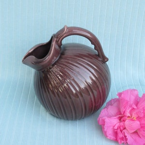 May include: A dark purple ceramic pitcher with a swirled design and a handle. The pitcher is sitting on a light blue surface with a pink flower.