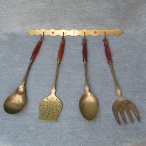 May include: A set of four vintage brass kitchen utensils hanging from a decorative brass bar. The utensils include a spoon, slotted spatula, solid spoon, and fork, each with a wooden handle. The brass has a warm, aged patina.