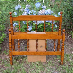 May include: A wooden bed frame with a brown finish and a spindle design. The frame is in a grassy area with a bush of blue flowers in the background.