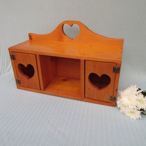 May include: A wooden wall shelf with two doors and a center open shelf. The doors and top of the shelf have heart-shaped cutouts. The shelf is stained a light orange color.