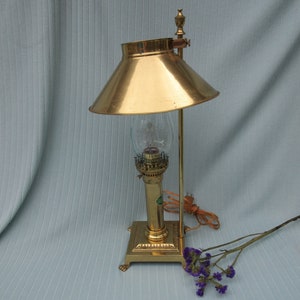 May include: A gold-toned table lamp with a glass shade and a square base. The lamp has a decorative finial on top and a cord with a plug.