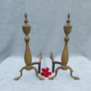 May include: A pair of antique brass fireplace andirons with a decorative finial on top. The andirons have three legs and are in good condition.