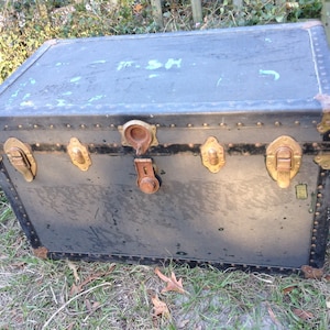 May include: A vintage black metal trunk with brass hardware and a worn finish. The trunk has a latch, two clasps, and a keyhole. The top of the trunk has a faded inscription that says "F.S.H."