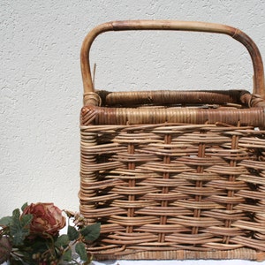 May include: A rectangular, woven wicker basket with a curved handle. The basket is a natural brown color and features a tightly woven pattern. A dried rose and greenery are in the foreground, suggesting a rustic or farmhouse theme.