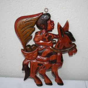 May include: A wooden carving of a person riding a donkey. The person is wearing a patterned shirt and has a dark brown hat. The donkey is brown and has a black mane. The carving is painted in shades of brown, red, and black.