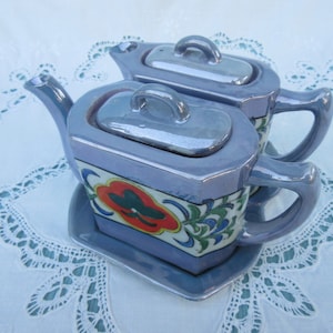 May include: A set of two vintage teapots with lids, featuring a floral design in shades of blue, green, red, and yellow on a lavender background. The teapots have silver-colored lids and handles.